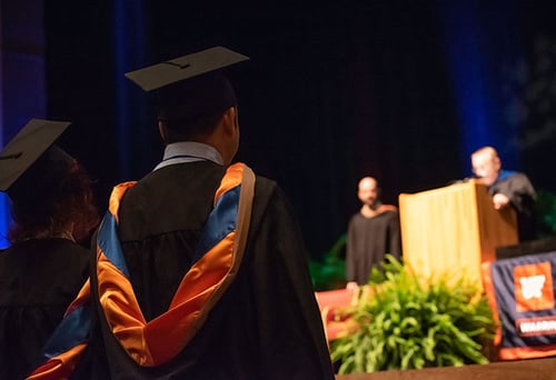 Isang larawan ng isang graduation event ang nakatuon sa dalawang estudyanteng nakikinig sa kanilang mga guro sa entablado.